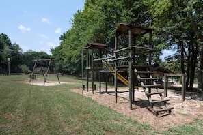 A playground with wooden structures, including a swing set and climbing frames, surrounded by trees.