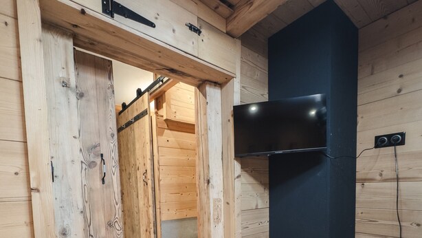 Ett träinredat utrymme med en skjutdörr som leder till ett badrum, en väggmonterad TV och ett eluttag på väggen.