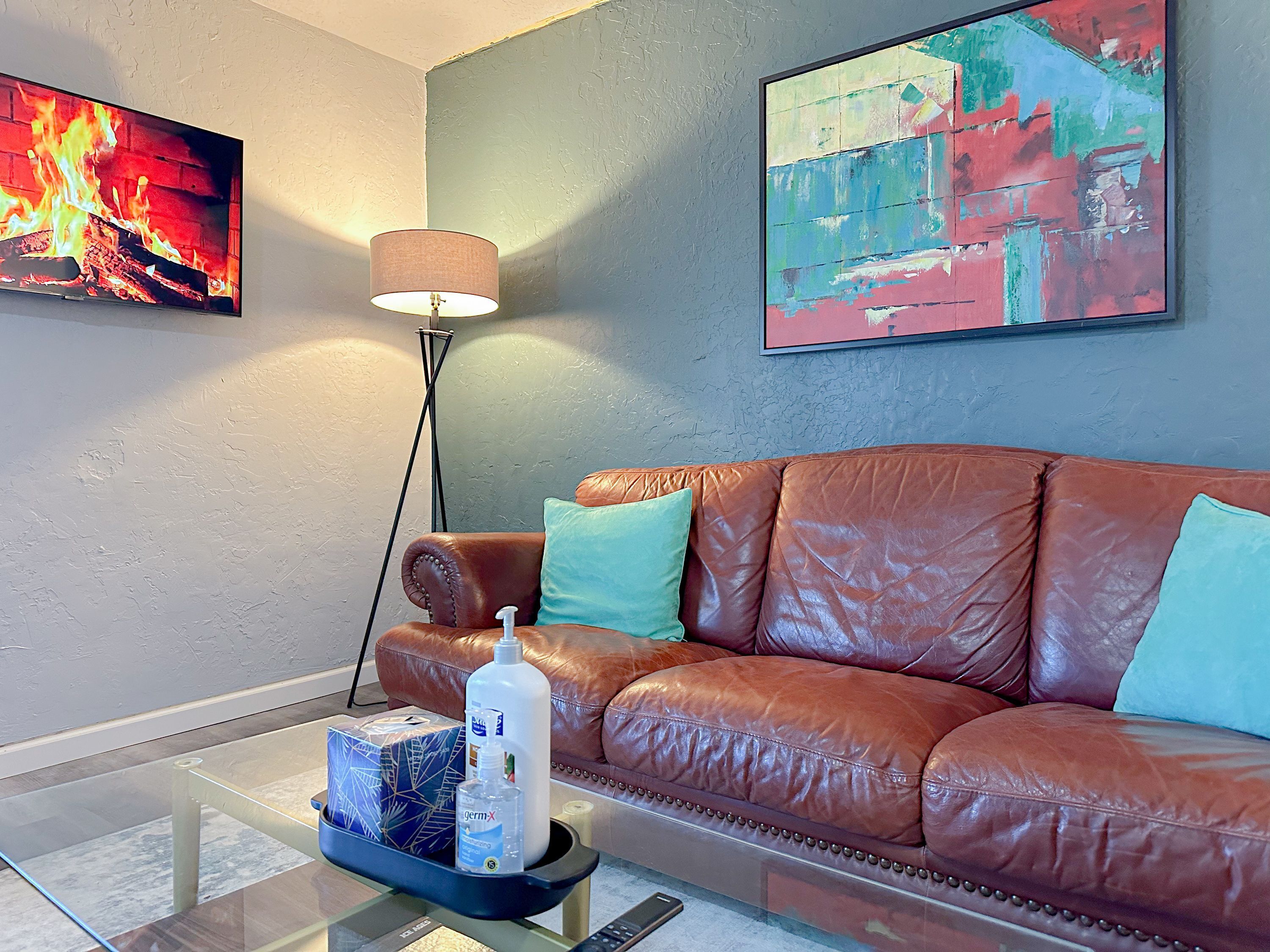 A living room with a brown leather sofa, a glass coffee table with a bottle of hand sanitizer, a lamp, and two paintings on the wall.