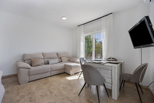 Un salon moderne comprenant un canapé, une table à manger, des chaises, un téléviseur à écran plat et une fenêtre avec des rideaux.