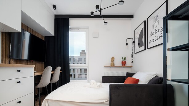 A modern hotel room with a bed, a desk with two chairs, a TV, a window with a view of buildings, and a shelf with decorative items. - Platon 46 Modern Studio (Łódź)