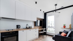 A modern kitchen with white cabinets, a wooden countertop, and a built-in oven. There is a dining area with a black chair and a black sofa with a red pillow. A window provides a view of buildings outside. - Platon 46 Modern Studio (Łódź)