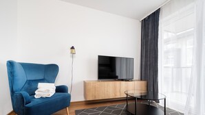 Una habitación de hotel moderna con un sofá azul, un televisor de pantalla plana sobre un mueble de madera, una mesa de centro de vidrio y un ventanal con cortinas translúcidas.