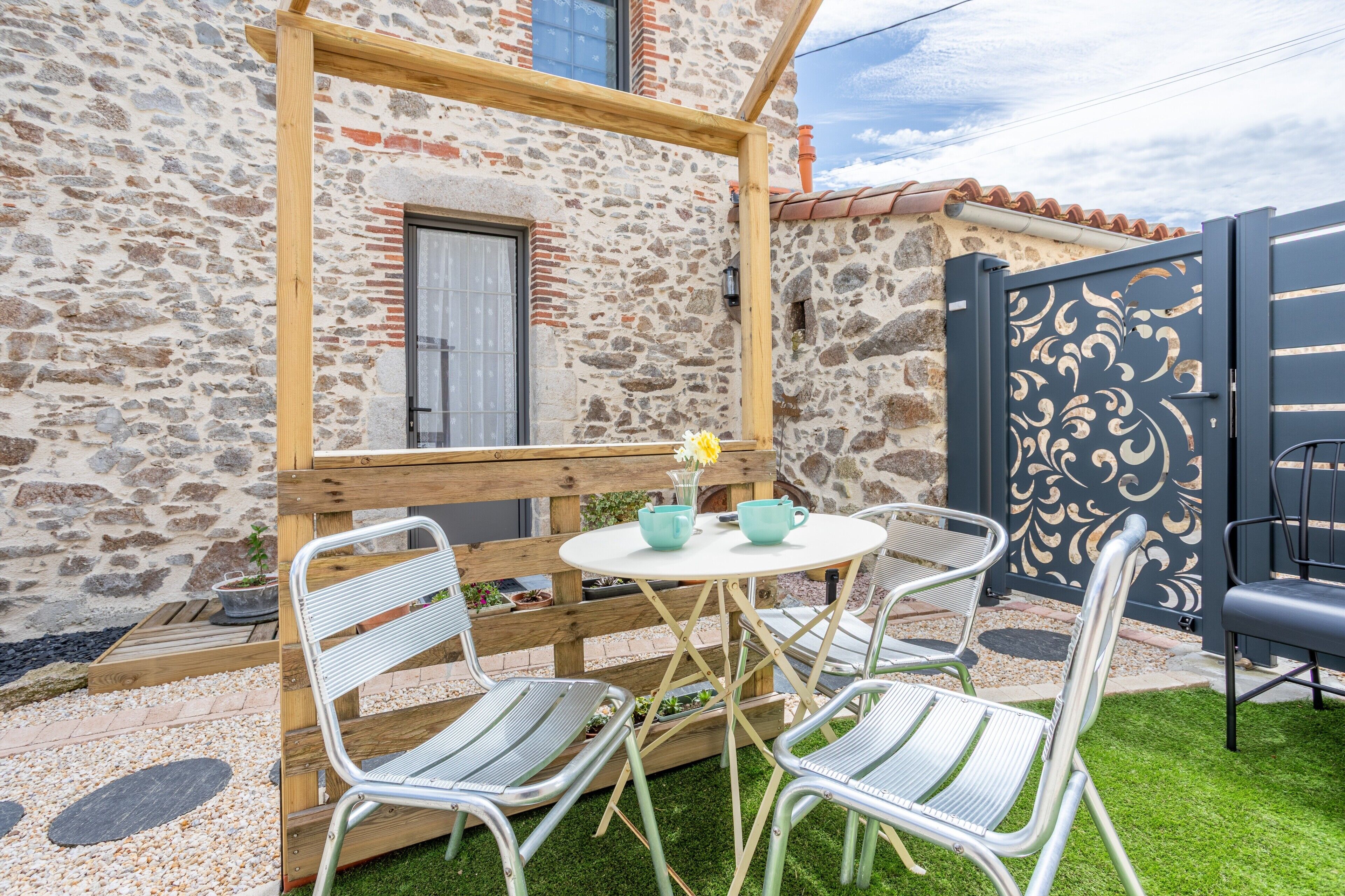 A stone building with a wooden pergola, a round table with chairs, and a decorative gate.