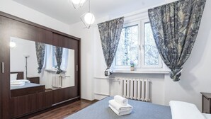 Een slaapkamer met een grote spiegel, een bed met een blauwe plaid, een houten garderobe en een raam met bloemen gordijnen.