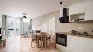 A modern kitchen with a built-in oven, wooden dining table, and chairs, a sofa, and a large window with curtains.