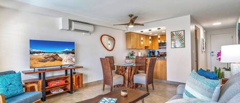 Uma sala de estar moderna com televisão de tela plana, mobiliário de madeira e uma área de refeições, com cozinha ao fundo.