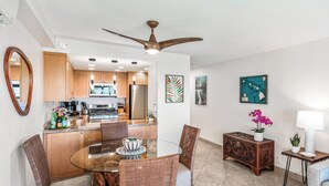 A modern kitchen with wooden cabinets, a stainless steel refrigerator, and a dining area with wicker chairs and a glass table. - Modern & Central Oceanview Vistas (Kihei)