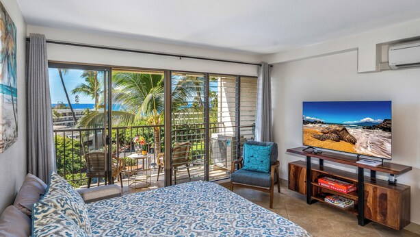 A hotel room with a large bed, a TV, and a balcony with a view of the ocean and palm trees. - Modern & Central Oceanview Vistas (Kihei)