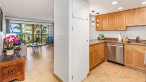A modern kitchen with wooden cabinets, a stainless steel dishwasher, and a large window overlooking a balcony with tropical plants. - Modern & Central Oceanview Vistas (Kihei)