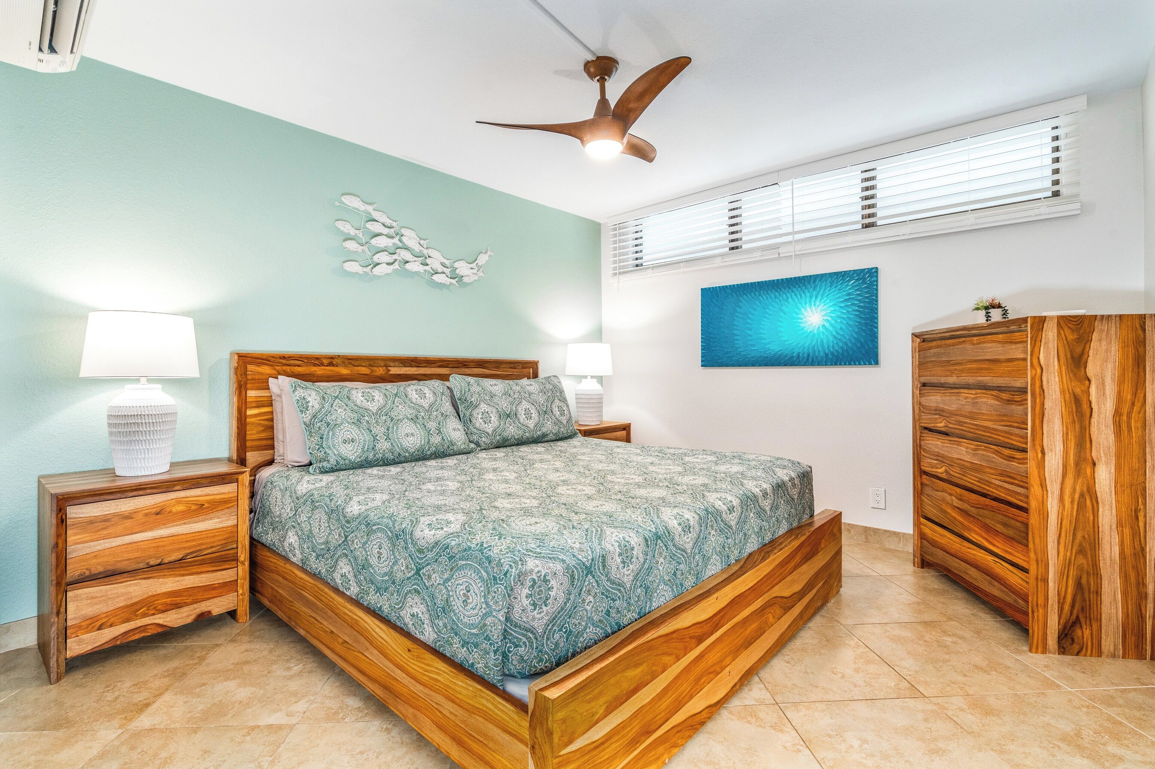 A bedroom with a wooden bed, two bedside tables with lamps, a ceiling fan, a wall-mounted air conditioning unit, a decorative wall piece, and a large window with blinds.
