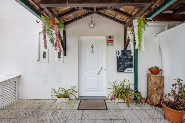 Una puerta blanca con felpudo, plantas colgantes y un cartel.
