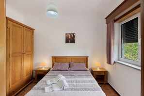 Un dormitorio con un armario de madera, una cama con cabecera, dos mesitas de noche con lámparas, una ventana con cortinas y un cuadro colgado en la pared.