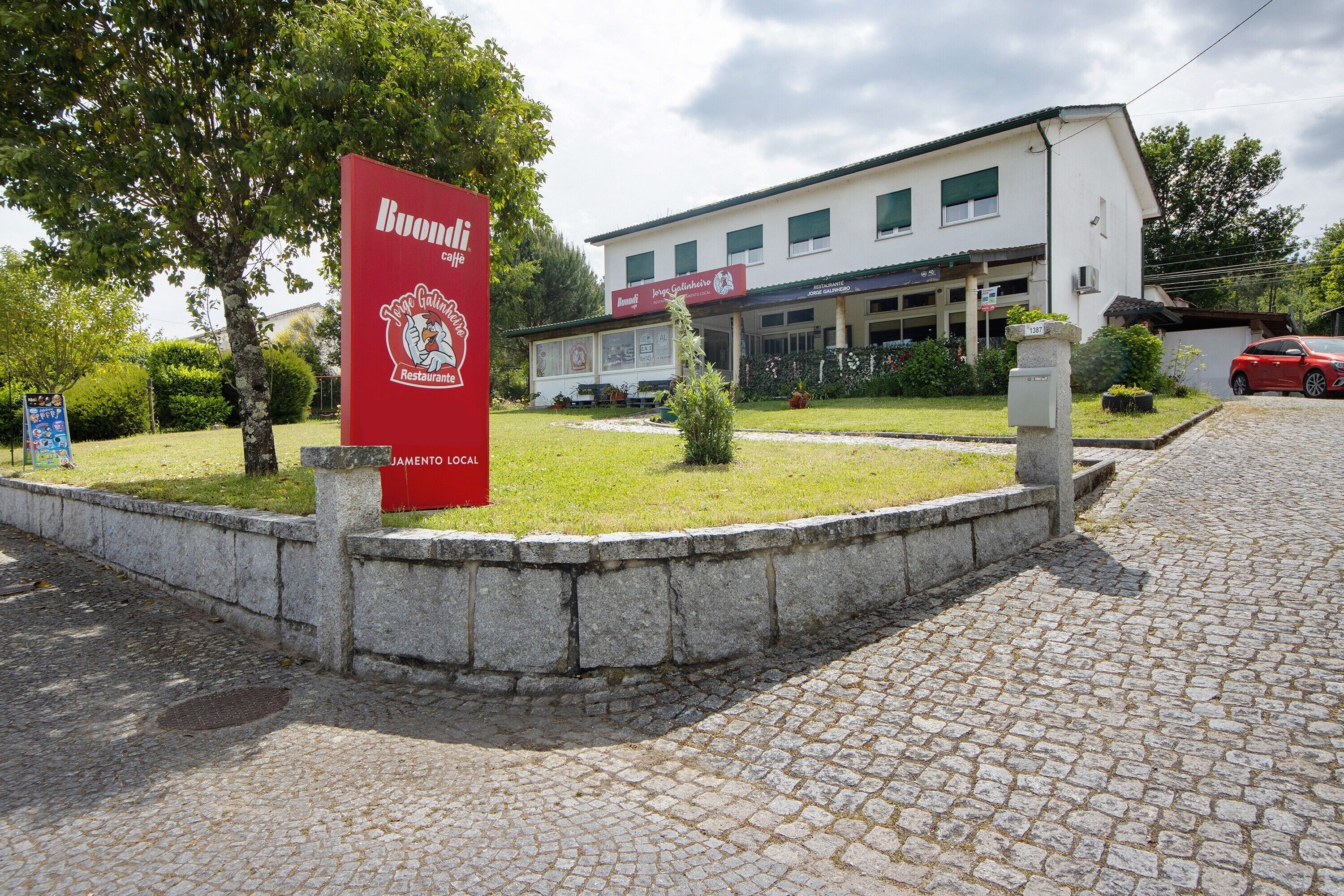 A building with a signboard, a paved area, and a red car parked nearby.