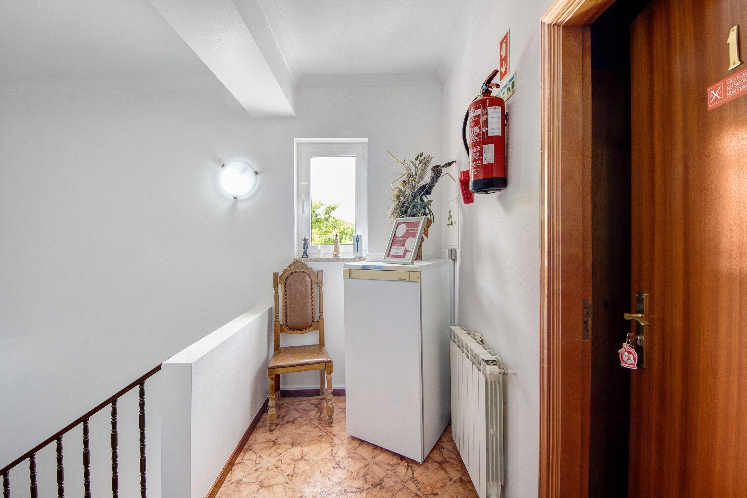 Un pasillo con una silla de madera, un electrodoméstico blanco, un extintor y una puerta con un número.