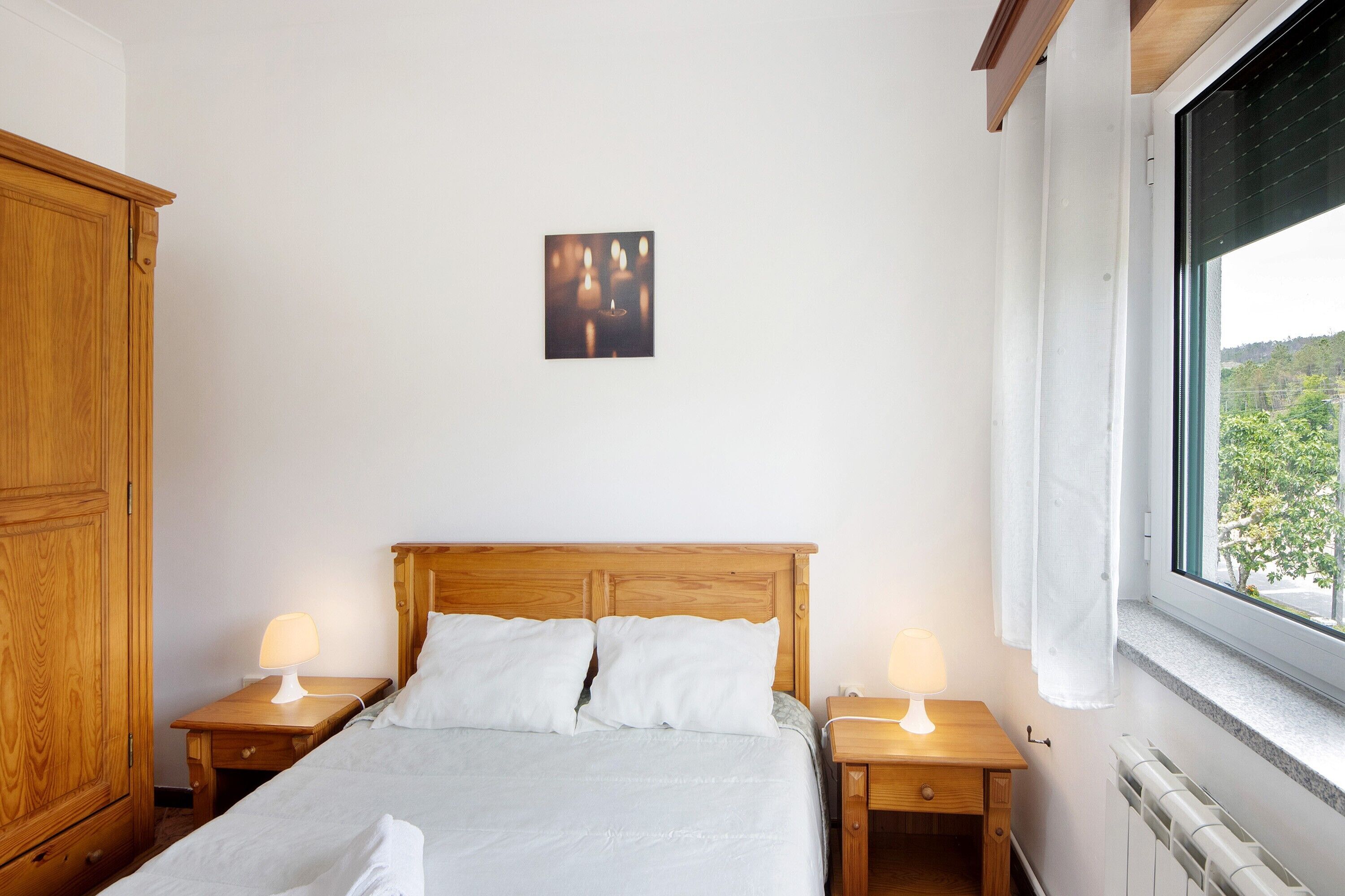 A bedroom with a wooden bed, two bedside tables with lamps, a radiator, and a window with a view.