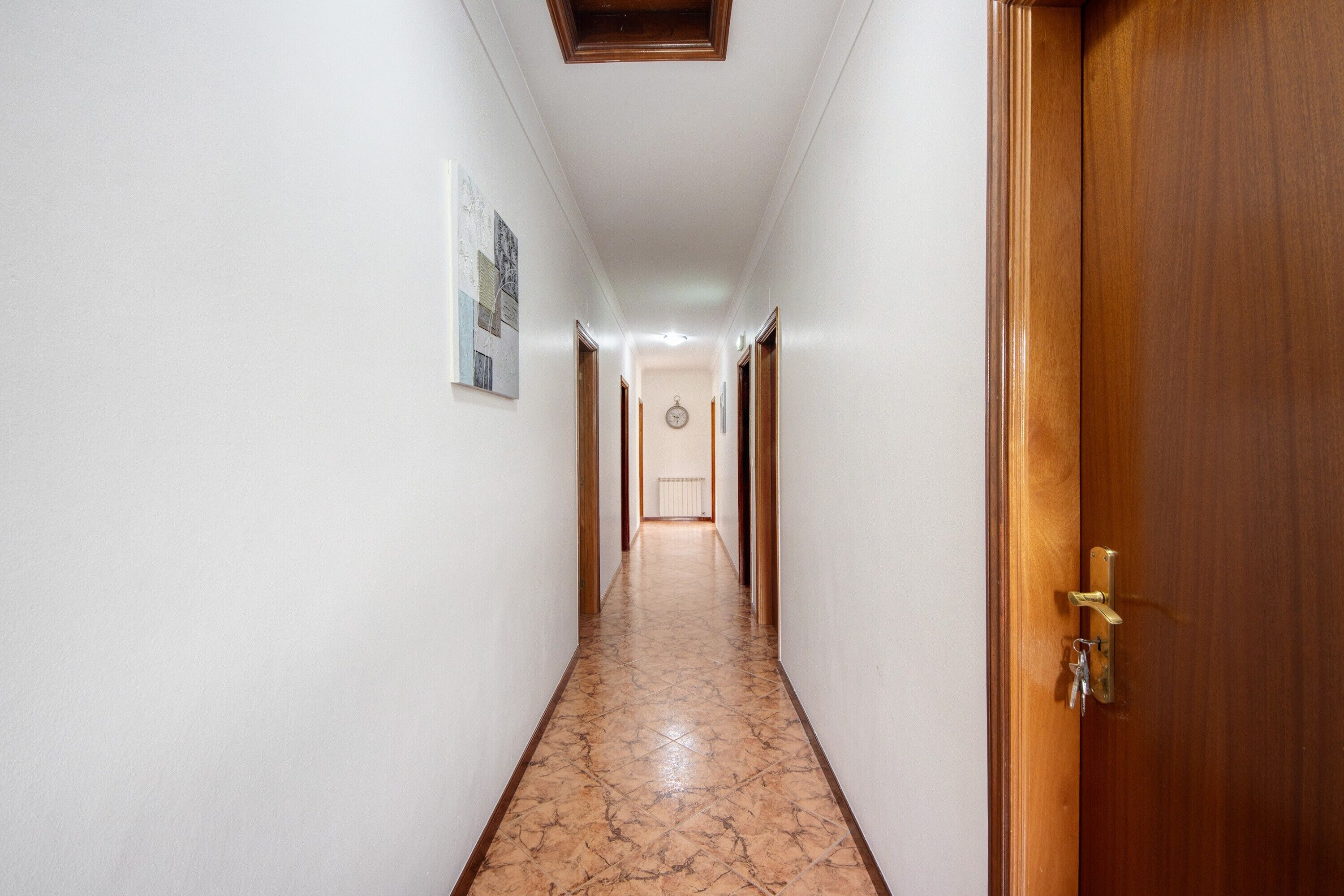 A hallway with a wooden door on the right, a tiled floor, and a clock on the wall.