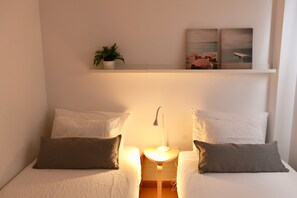 A bedroom with two beds, each with a pillow and a bedside table with a lamp. There is a shelf above the beds with a plant and two framed pictures.