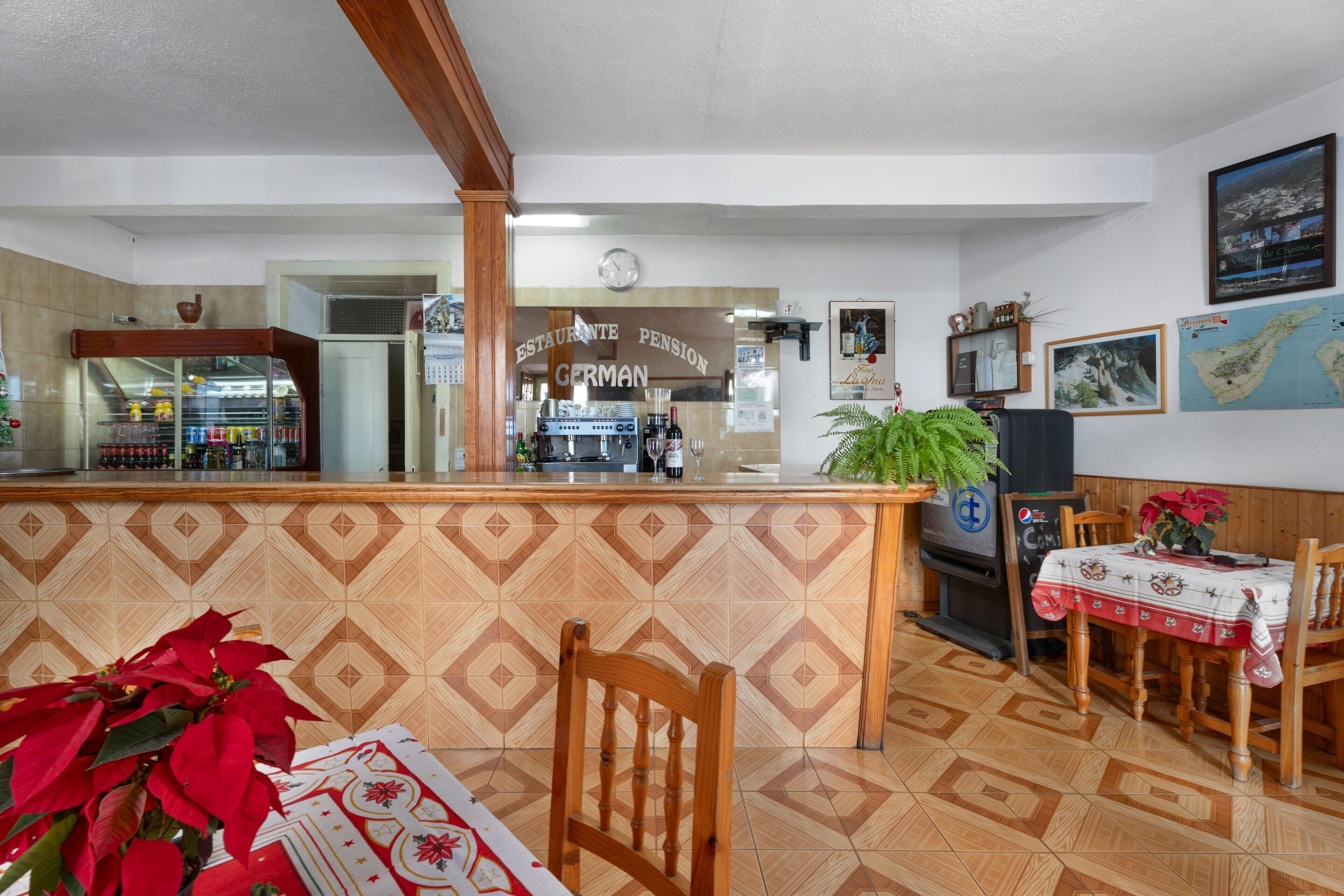 A restaurant with a counter, wooden chairs, a table with a tablecloth, a clock on the wall, and a menu board.