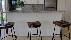 Une cuisine moderne avec des armoires blanches, un comptoir en granit, un réfrigérateur en acier inoxydable, un four à micro-ondes et un évier. On y trouve trois tabourets de bar avec des pieds en métal et une plante en pot sur le comptoir.