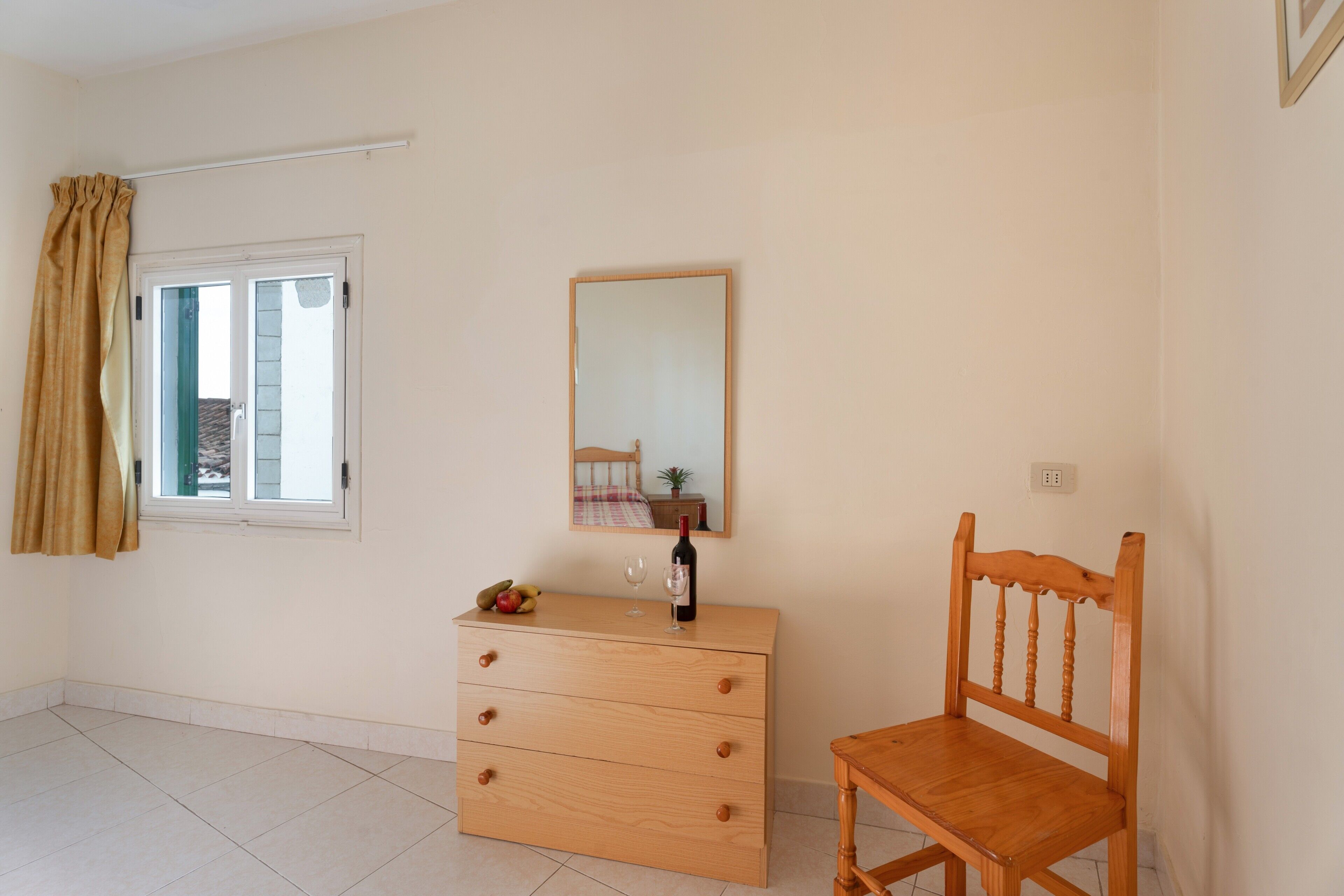 Una habitación con un aparador de madera, una silla, un ventanal con cortinas, un espejo y piso de baldosas.