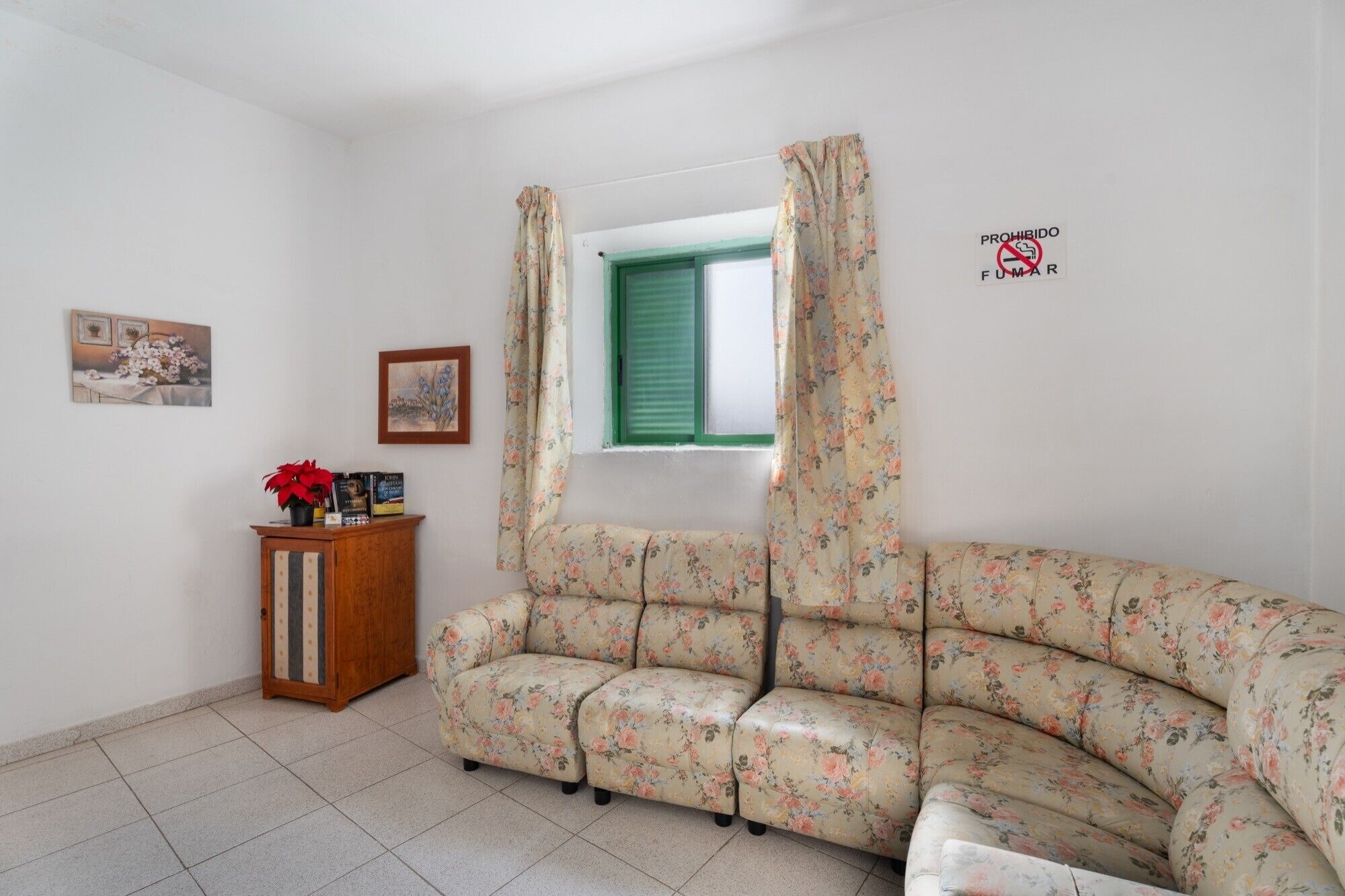 Een woonkamer met een bloemenbank, een houten kast, een raam met groene luiken en een 'No Smoking' bordje.