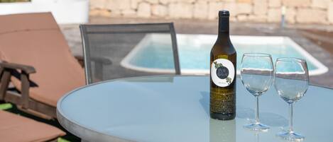 Ein runder Glastisch mit einer Flasche und zwei Gläsern, ein Stuhl und ein Pool im Hintergrund.