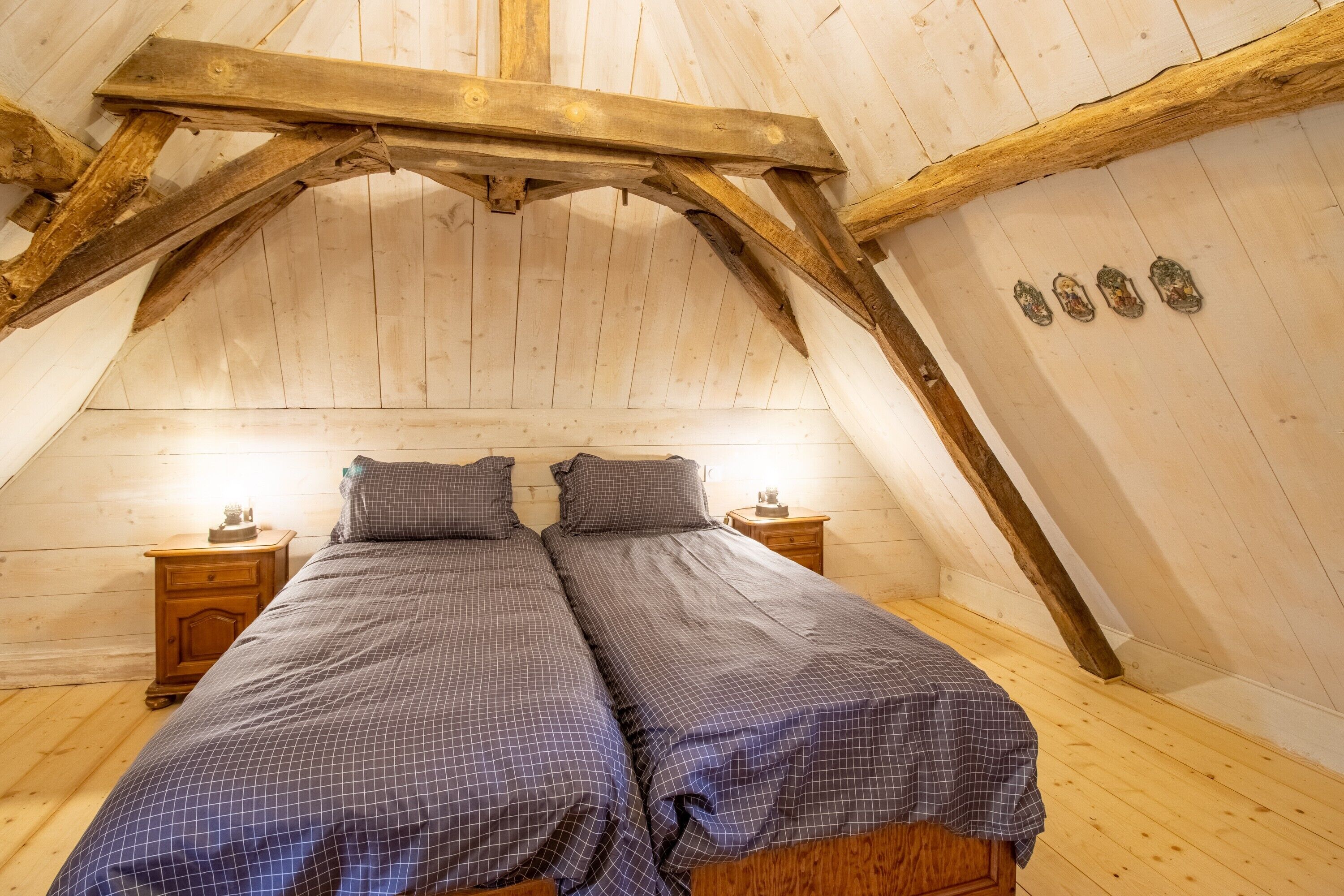 Une pièce avec un plafond en pente, des poutres en bois, deux lits avec une literie à carreaux, des tables de chevet avec des lampes et un plancher en bois.