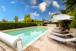 Una piscina con lettini e ombrelloni, circondata da siepi e alberi.