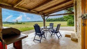 A covered patio with a grill, outdoor seating, and a view of a green field. - Chez Jacquou – Authentic Charm and Modern Comfort with Private Pool in the Heart of Périgord (Bouniagues)
