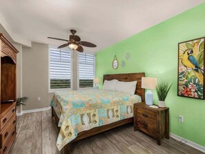 A bedroom with a bed, wooden nightstand, ceiling fan, and a green wall with a parrot painting. - Perfect Island Escape - Beach, Pool, Restaurant & More! -FMB-Condo Island Oasis - Roelens (Fort Myers Beach)