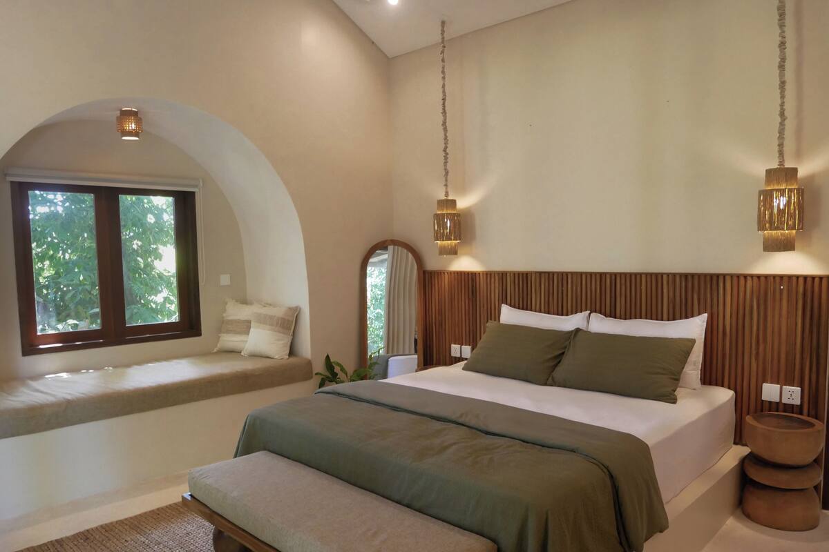 A bedroom with a large bed, a bench at the window, and a view of trees outside.