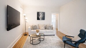 Un soggiorno moderno con una televisione a schermo piatto, un divano con cuscini decorati, una poltrona blu, una lampada da terra e un quadro appeso al muro.