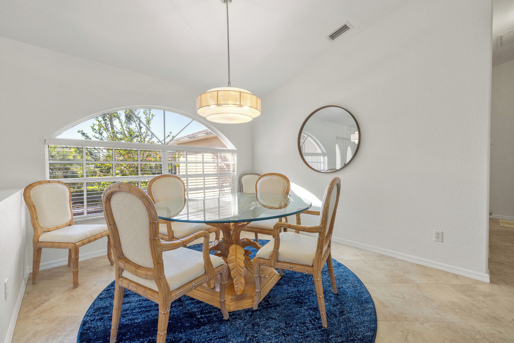 Un comedor con mesa de vidrio rodeada por cuatro sillas, alfombra azul, lámpara colgante y espejo redondo en la pared.