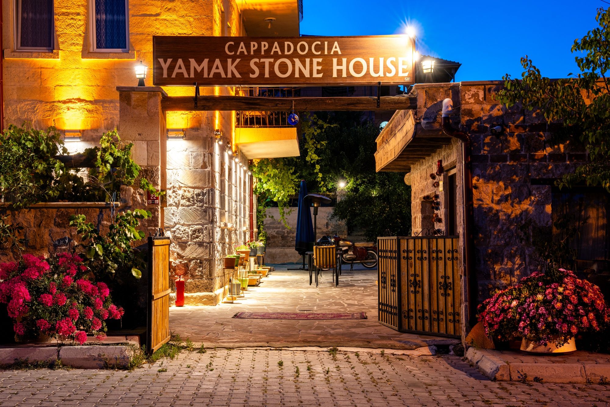 La entrada a Cappadocia Yamak Stone House, con fachada de piedra, un letrero de madera y macetas con flores.