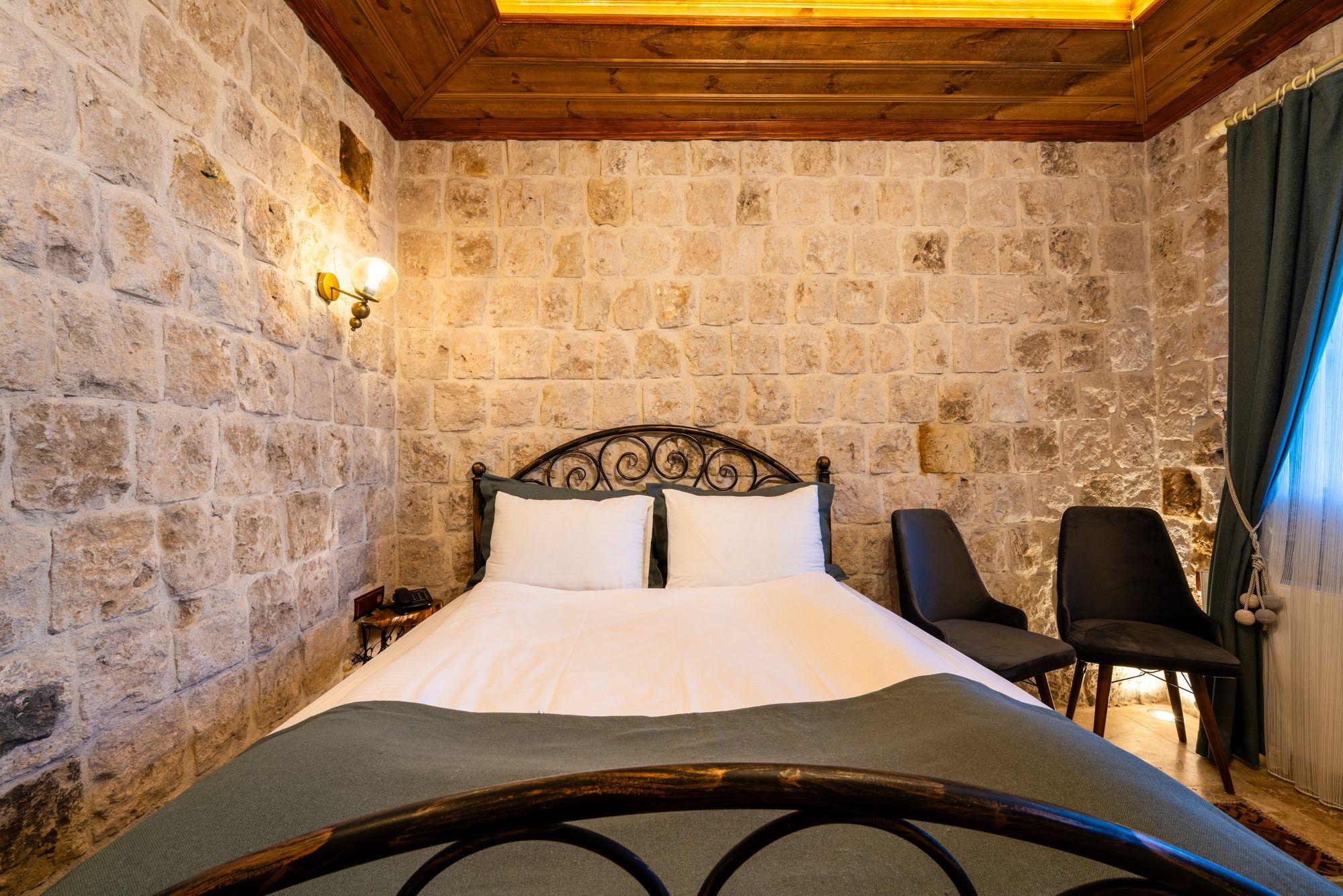A room with a stone wall, a wooden ceiling, a bed with white bedding, two chairs, and a nightstand with a lamp.