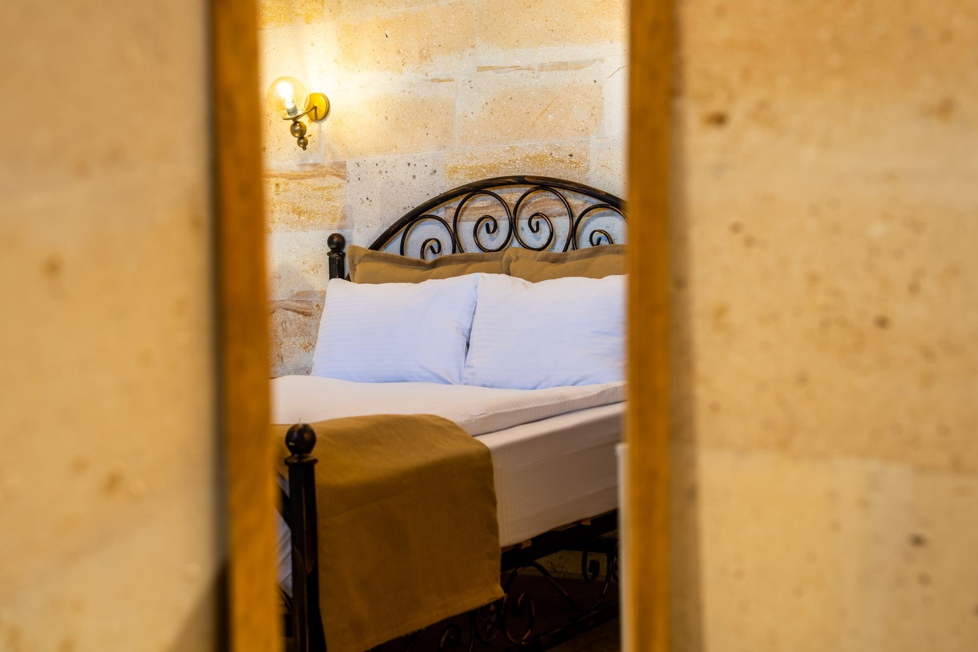 Una cama con cabecera, un aplique de pared y una vista parcial de una habitación con pared de piedra.