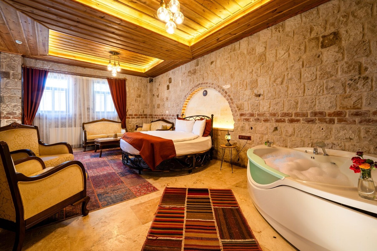 A hotel room with a bed, a sitting area with a sofa and armchairs, a large bathtub, and a stone wall.
