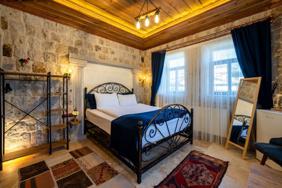 A bedroom with a stone wall, a bed with a metal frame, a wooden ceiling, and a large window with curtains.