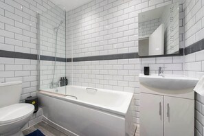 Une salle de bain moderne avec des carreaux de céramique blancs rectangulaires, une douche à parois de verre, une baignoire blanche et un lavabo avec un miroir au-dessus.