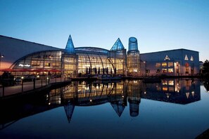 Een modern gebouw met een glazen gevel, een reflecterend waterlichaam ervoor en een brug die verschillende delen verbindt.