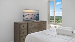 Een slaapkamer met een groot bed, een dressoir, een flatscreen televisie en een raam met uitzicht op groen en gebouwen.