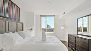 A modern bedroom with a large bed, a dresser, a window with a city view, and a framed artwork above the bed. - Tysons 2BR | Panoramic Views, Rooftop Pool & Gym (McLean)