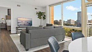 A modern living room with a large window, a grey sofa, a dining table, and a TV. - Tysons 2BR | Panoramic Views, Rooftop Pool & Gym (McLean)