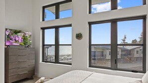 A bedroom with a bed, a dresser, and a TV on a dresser. There are large windows with a view of a beach and buildings. - Sea and Ski at The Beach House (Black Creek)
