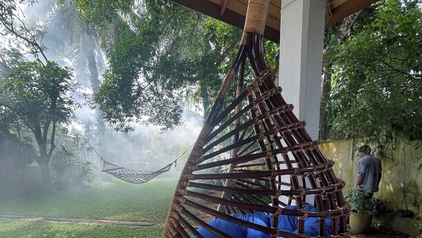 Un fauteuil suspendu avec des coussins bleus, un sol carrelé et une vue sur un jardin avec des arbres et un hamac au fond.