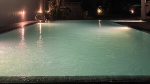 Une piscine éclairée la nuit, avec un arbre en arrière-plan et une zone abritée sur la droite.