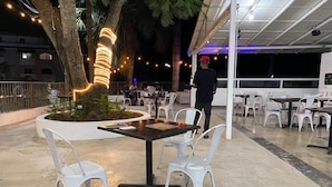 An outdoor dining area with tables and chairs, surrounded by palm trees and string lights.