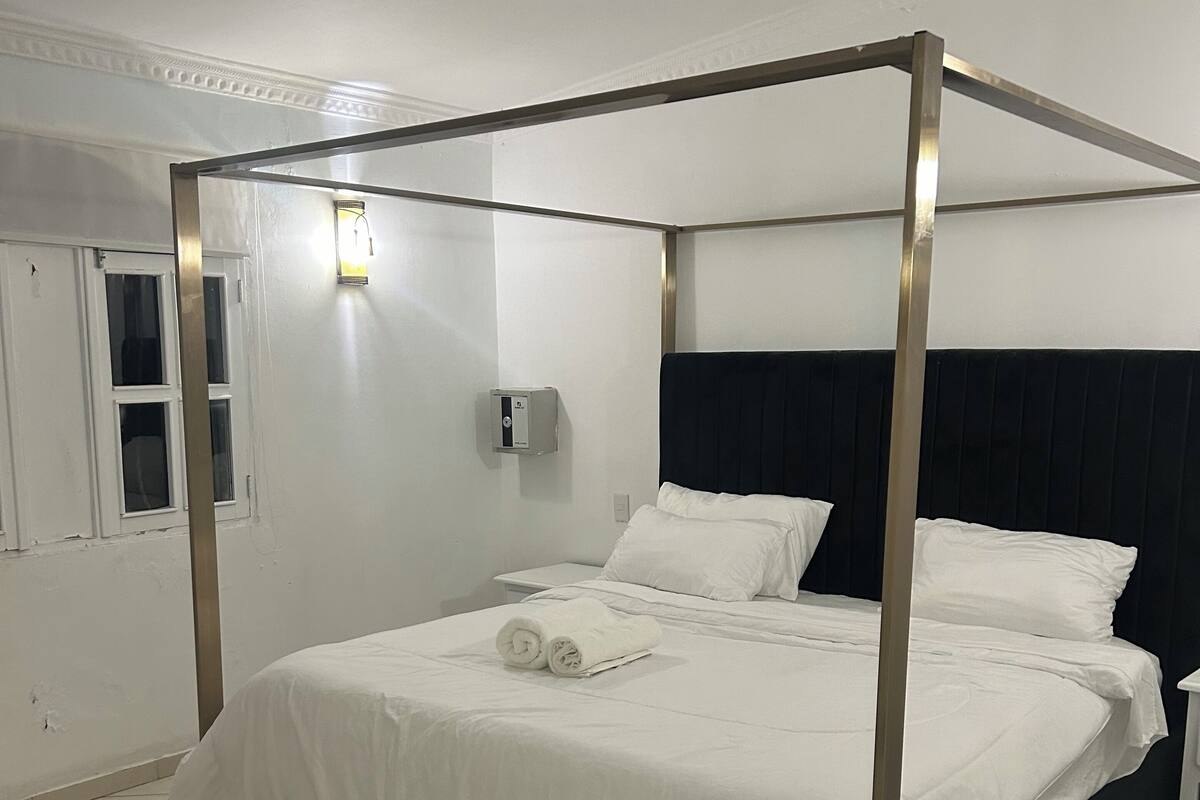 Ein Bett mit Baldachin, ausgestattet mit weißen Leinen und einem schwarzen Kopfteil, ein Nachttisch, eine Wandlampe und ein Fenster mit Jalousien.
