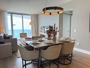 Een moderne eethoek met een grote tafel gedekt voor vier personen, omringd door stoelen. Een kroonluchter hangt boven de tafel, en er staan twee vazen met bloemen op de tafel.